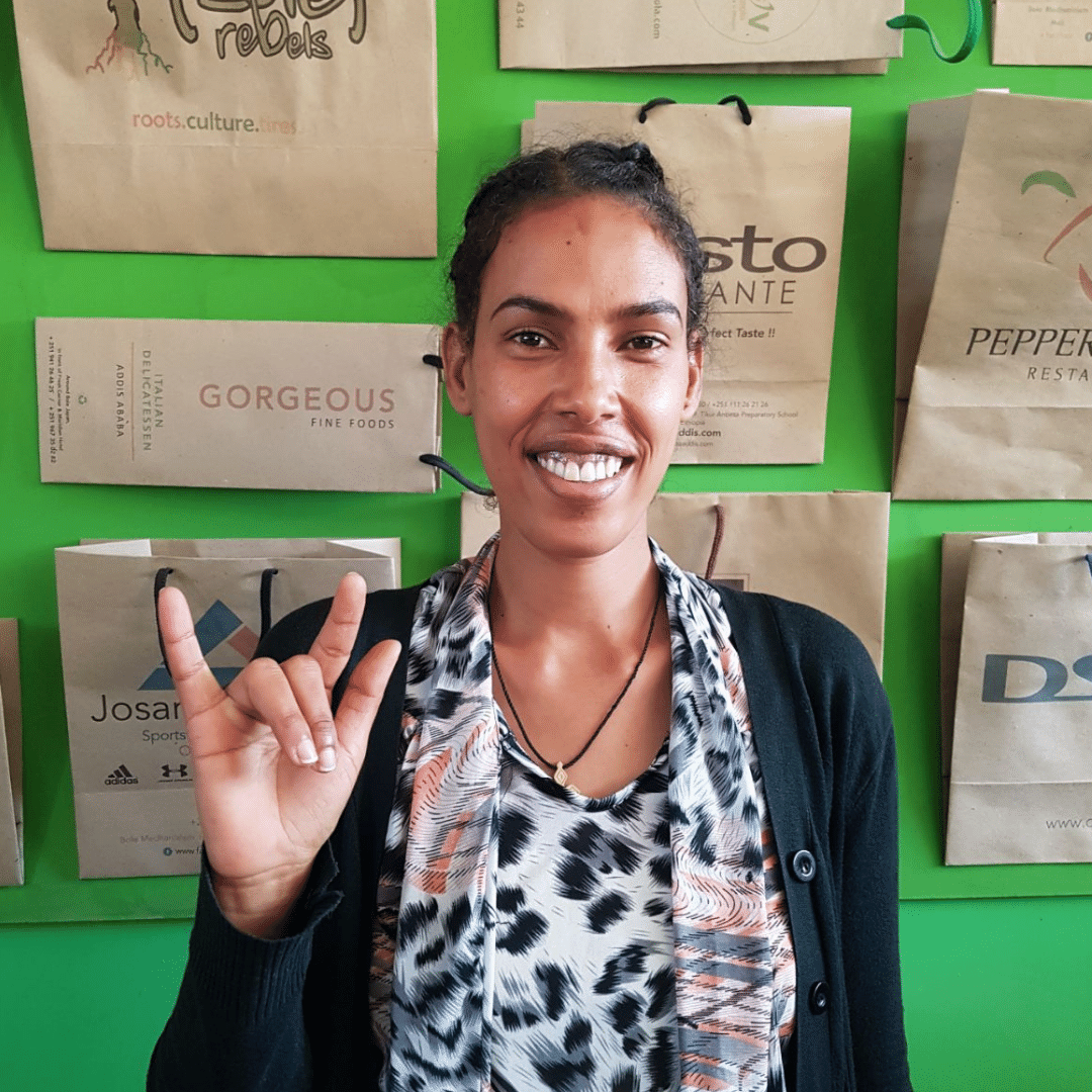 A picture of Nigist one of the Teki Paper Bags employees who is deaf. She is wearing a black jacket and pose infront of a green wall.